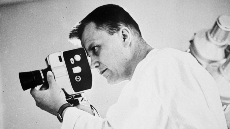 Black-and-white image of a man filming with a handheld movie camera.