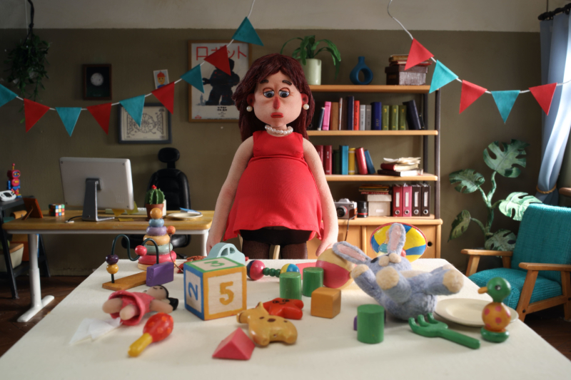 Leļļu animācijas aina, kurā grūtnieces lelle stāv pie galda ar bērnu rotaļlietām, fonā redzams mājīgs interjers ar grāmatplauktu un svētku karodziņiem.