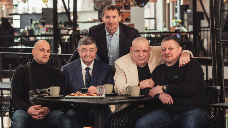 Pieci vīrieši pozē ap galdu kafejnīcā.