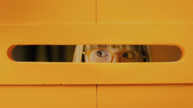 Caur spaugu oranžā sienā redzamas bērna acis ar brillēm, raugāmies sānis.