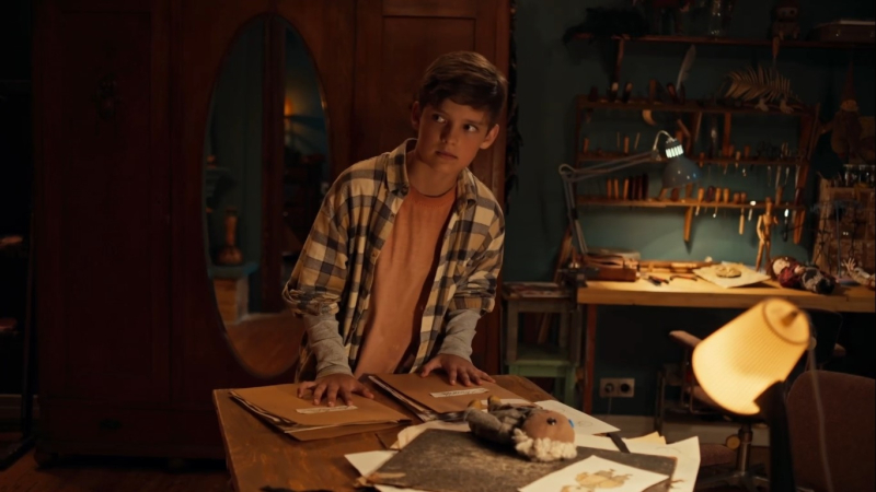 Zēns stāv pie darba galda mājīgā darbnīcas telpā un ar piesardzīgu skatienu raugās sānis.