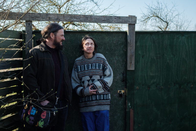 Pie zaļiem dārza vārtiem stāv un viens uz otru sāņus lūkojas vīrietis un sieviete.