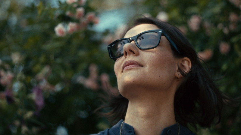 Jauna sieviete saulesbrillēs skatās augšup uz ziedu fona.