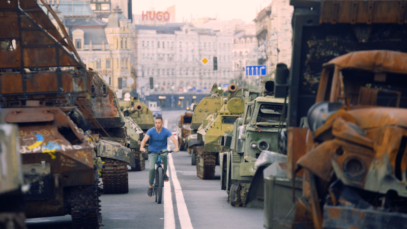 Zēns ar velosipēdu brauc caur ielas vidū izliktiem iznīcinātiem militāriem transportlīdzekļiem.