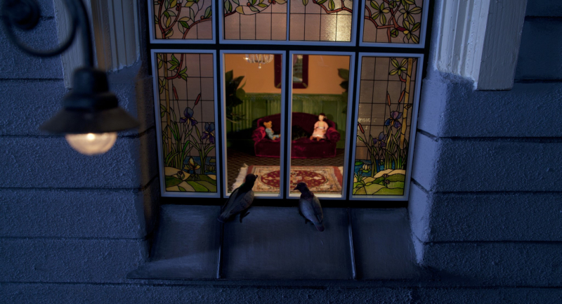 Skats caur vitrāžas logu uz mājīgu istabu ar lellēm uz dīvāna.