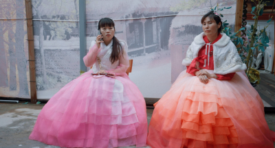 Blakus sēž divas sievietes krāšņos, kuplos svētku tērpos; viena runā pa telefonu.