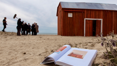 Priekšplānā smiltīs stāv atvērta grāmata, fonā sarkans koka šķūnis un filmēšanas komanda.
