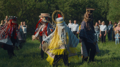  Cilvēki krāsainos, tradicionālos masku tērpos dejo pļavā skatītāju priekšā.