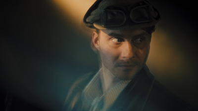 Tuvplāna portrets ar jaunu vīrieti tumšā cepurē, kinematogrāfiskā apgaismojumā.