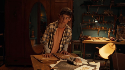 Zēns stāv pie darba galda mājīgā darbnīcas telpā un ar piesardzīgu skatienu raugās sānis.