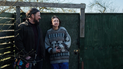 Two adults stand beside a weathered green wooden gate outdoors; the man looks at the woman while she gazes upward with a thoughtful expression.
