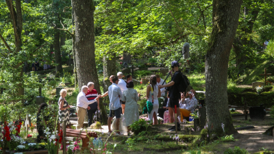 Cilvēku grupa pulcējusies kapsētā starp kokiem, daļa sarunājas pie kapavietām, redzams arī filmēšanas komandas dalībnieks ar mikrofonu.